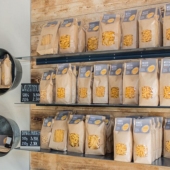 Rechts sind drei Regalbretter an einer Holzwand angebracht. Darauf stehen mehrere Nudelpackungen. Daneben an einer weißen Wand sind verschieden große Zinswannen mit Innenbrett als Regale angebracht, in denen sich Regionale Produkte wie Eierlikör, Eier und Nudelpackungen befinden.