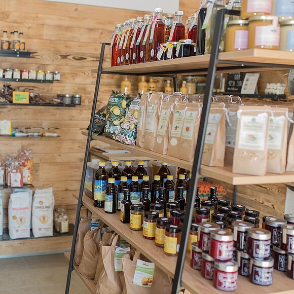 Von der Seite aufgenommen sieht man rechts ein großes Regal mit Eisenträgern und 3 langen Holzbrettern gefüllt mit verschiedenen regionalen Produkten wie Marmeladen, Eßige, Öle, Papiertüten mit Kartoffeln ganz unten, Müsli, Chips, Honig. Im Hintergrund an der Holzwand sind Regalbretter befestigt, auf welchen sich noch weitere Produkte wie Papiertüten mit Mehl, Kräutersalze und Eierlikör befinden.