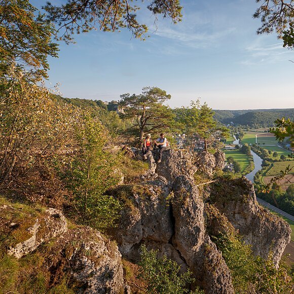 Wanderer auf der Arnsberger Leite am Altmühltal-Panoramaweg Ein Wanderpärchen, etwas entfernt, steht links auf einem Felsen an der Arnsberger Leite, die gut zu sehen ist. Dahinter erstreckt sich der Blick ins Tal. Der Himmel erstrahlt wolkenlos.