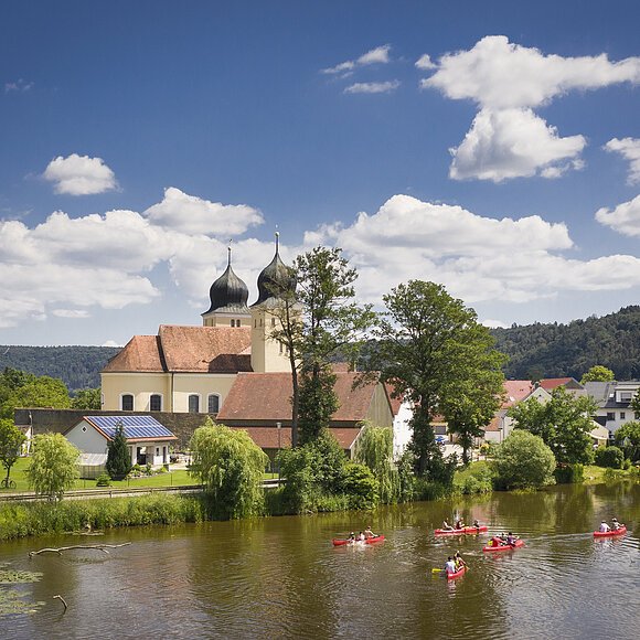 Auf der Altmühl paddeln fünf rote Kanus an Kottingwörth mit ihrer Kirche und deren zwei Kirchtürmen vorbei.