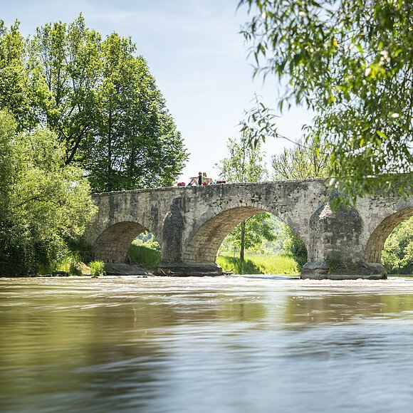Römerbrücke Pfünz Auf der Römerbrücke sitzen zwei Wanderer Rücken an Rücken auf dem Steingeländer der Brücke. Eingerahmt von Bäumen und unter ihnen die Altmühl.