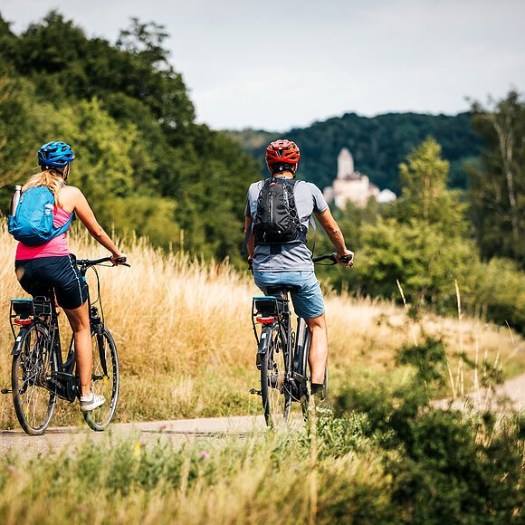 Zwei Radler, eine Frau und ein Mann, sind auf dem Radweg unterwegs. Die Gräser entlang des Radweges sind recht schon recht vertrocknet und braun. Ihr Ziel ist die Burg Kipfenberg die inmitten des Waldes in weiß bereits unscharf zu sehen ist.