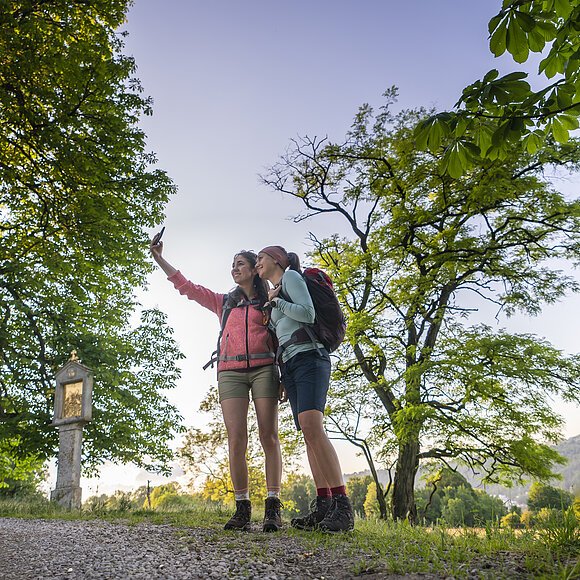 Zwei Frauen in rosafarbenen und hellblauem Shirt, sowie kurzen Hosen stehen eng beieinander am Frauenberg und die Frau in rosa, hält ihr Handy für ein gemeinsames Selfie in die Höhe. Ein großer Baum, sowie ein Marterl sind vor den Zweien zu erkennen. Im Hintergrund der zwei Frauen sind vereinzelt Bäume zu sehen , aber auch ein wolkenloser Himmel.