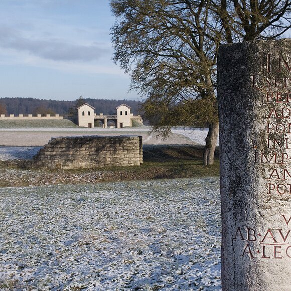 Im Vordergrund befindet sich ein römischer Meilenstein mit Entfernungsangaben zu den nächsten wichtigen Zielen. Im Hintergrund der winterlichen Landschaft ist die rekonstruierte Toranlage des römischen Kastells Vetoniana zu sehen.