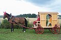 Ein Pferd zieht einen hölzernen Wagen mit zwei Personen in historischer Kleidung über eine Wiese.