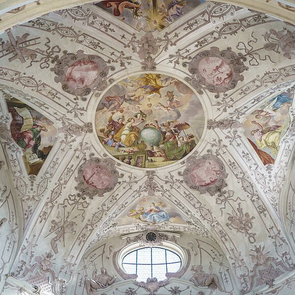 Deckenfresko mit religiösen Szenen und Ornamenten in einer Kirche, umgeben von Fenstern und Stuckverzierungen.