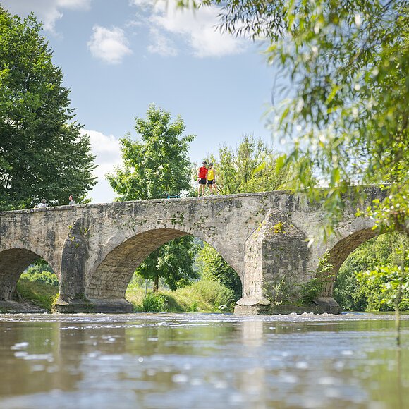 Steinbrücke mit drei Bögen über einen Fluss, umgeben von Bäumen und Vegetation.