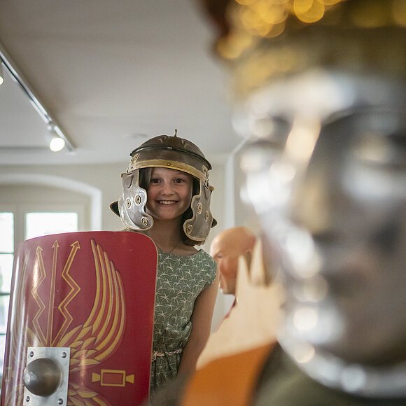 Kind trägt römischen Helm und hält Schild, im Hintergrund eine Statue.