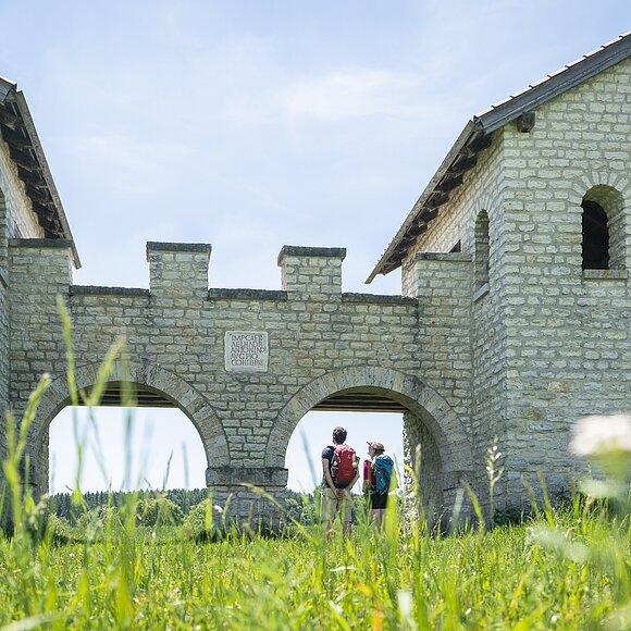 Römerkastell Vetoniana bei Pfünz Zwei Personen mit Rucksäcken stehen unter einem steinernen Torbogen einer historischen Anlage bei sonnigem Himmel.