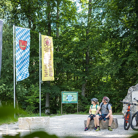 Zwei Radfahrer mit Helm sitzen auf Steinbank im Wald neben zwei Mountainbikes und drei Fahnen.