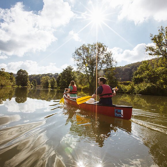 Zwei Personen paddeln in einem roten Kanu auf einem ruhigen Fluss bei sonnigem Himmel mit Wolken.