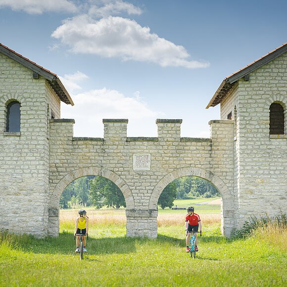 Zwei Radfahrer fahren durch zwei Bögen einer historischen Steinmauer in einer grünen Wiesenlandschaft.