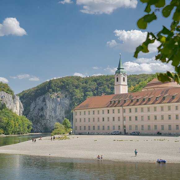 Flussufer mit Kiesstrand, altes Gebäude mit Turm und bewaldete Felsen unter blauem Himmel mit Wolken
