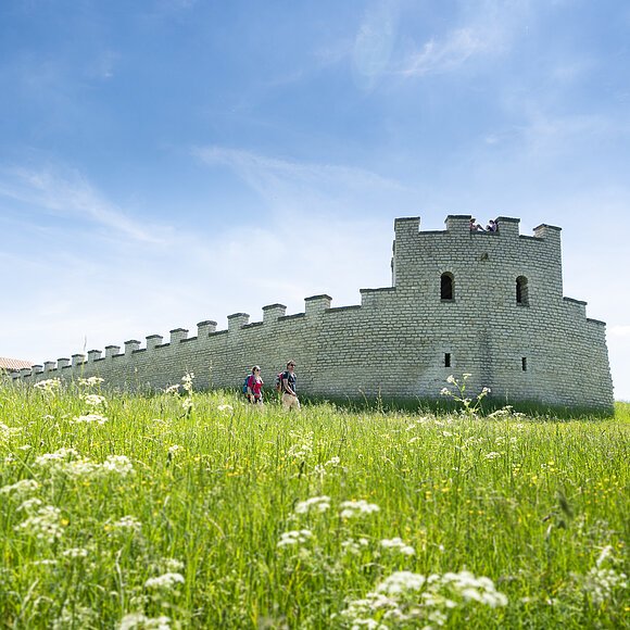 Zwei Wanderer gehen an einer historischen Steinmauer auf einer grünen Wiese unter blauem Himmel vorbei.