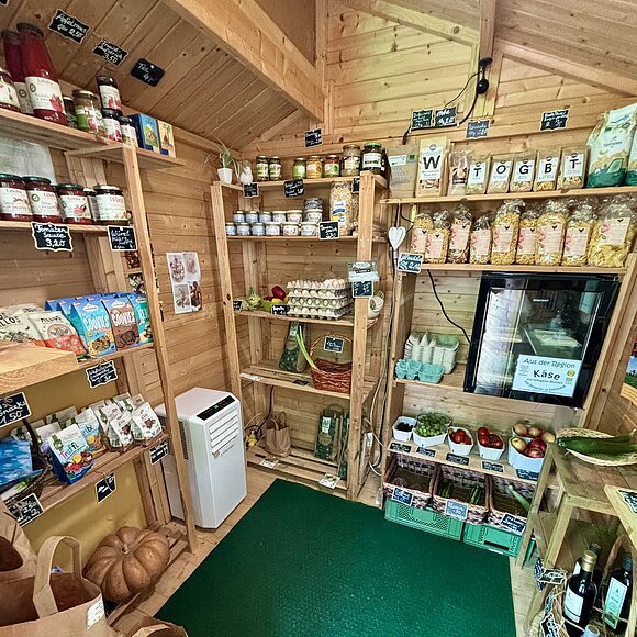Kleiner Holzladen mit Regalen voller Lebensmittel, Eiern, Obst, Gemüse und Getränken.