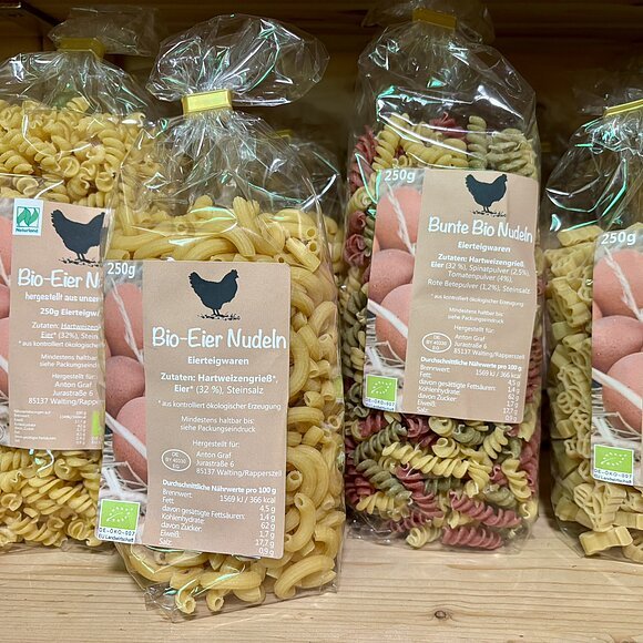 Verschiedene Packungen Bio-Eiernudeln in Plastikbeuteln auf Holzregal, Sorten und Formen sichtbar.