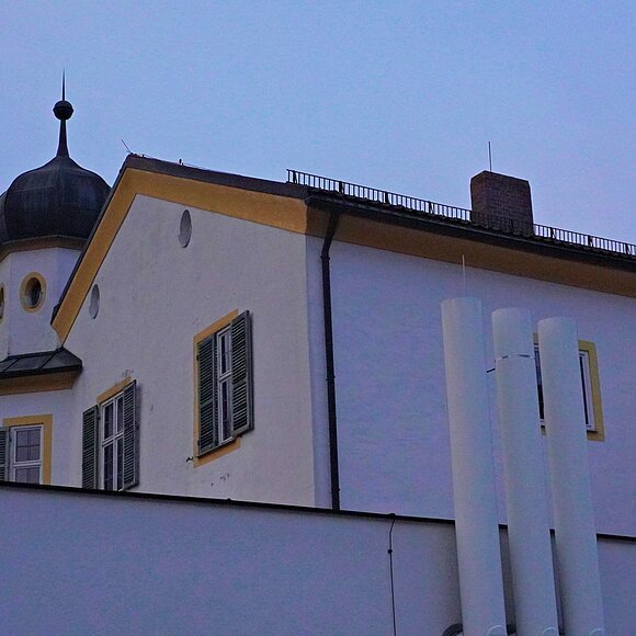 Walting Schloss Pfuenz Außenansicht Gebäude mit weißer Fassade, grünen Fensterläden und einem Turm mit schwarzer Kuppel bei Dämmerung.