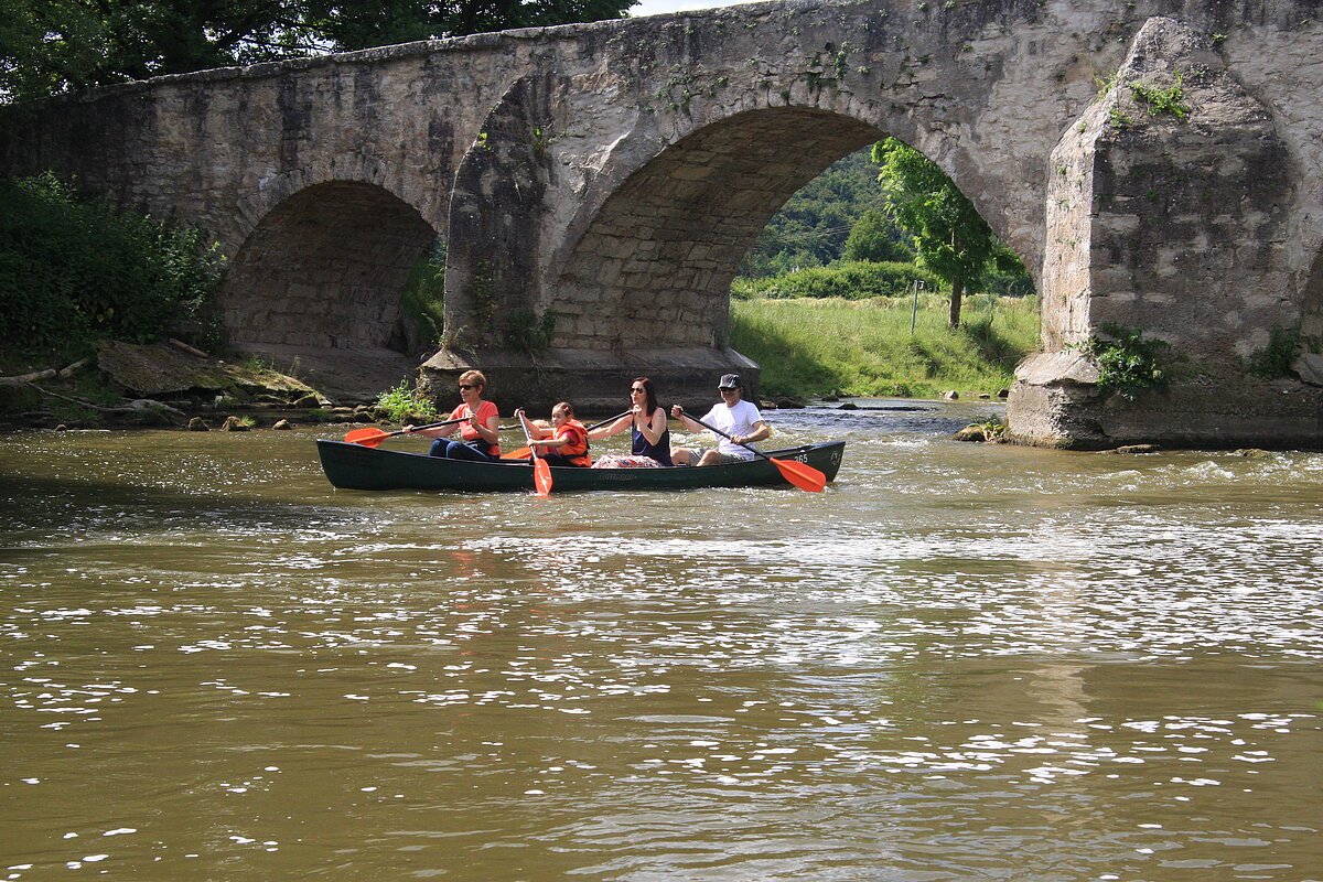 Eine Familie ist einem Kanu vom Schmidt Bootsverleih auf der Altmühl unterwegs. Sie sind gerade unter der Steinbrücke hindurchgefahren, diese ist in Teilen noch zu sehen.
