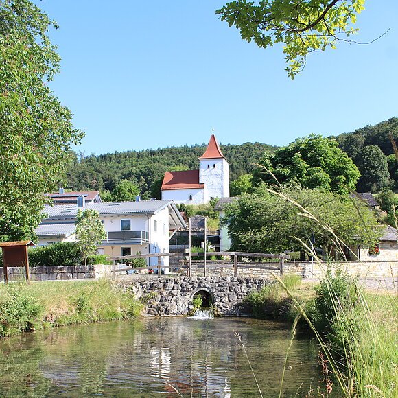 Fluss mit klarstem Wasser, Brücke aus Stein, Häuser und Kirche mit rotem Dach vor bewaldetem Hügel unter blauem Himmel.