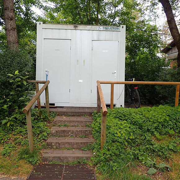 Bootsrastplatz Gungolding Außenansicht eines weißen Containers mit zwei Türen, beschriftet mit „Herren“ und „Damen“, umgeben von Grün und Bäumen.