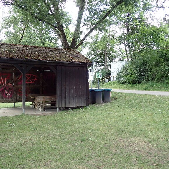 Bootsrastplatz Gungolding Holzunterstand mit Bank und Graffiti, daneben Mülltonnen und Waldweg im Grünen