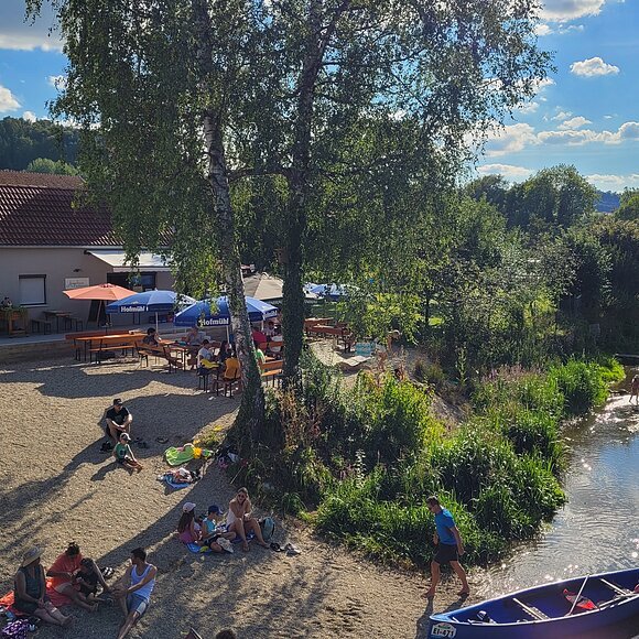 Angelinas Altmühlrast in Pfalzpaint Menschen sitzen am Ufer eines Flusses mit Kanus, Bäumen und einem Gebäude mit Sonnenschirmen im Hintergrund.