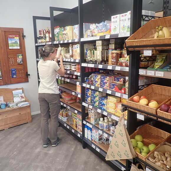 Gefüllte Regale im Dorfladen Heimat und Genuss Gungolding Frau steht vor einem Regal mit Lebensmitteln in einem kleinen Laden, rechts Obstkörbe mit Äpfeln und Bananen.