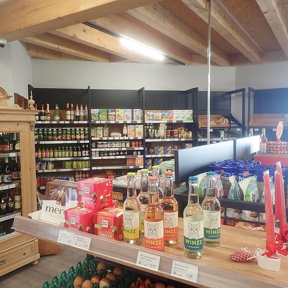 Sortiment des Dorfladen Heimat und Genuss Gungoldingheane Innenansicht eines kleinen Lebensmittelgeschäfts mit Regalen, Holzmöbeln und verschiedenen Produkten.